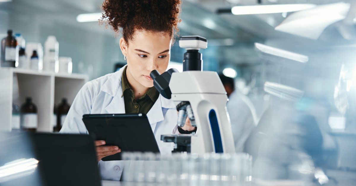 Female scientists wearing a white robe sitting behind a desk while holding a tablet and looking through a microscope.