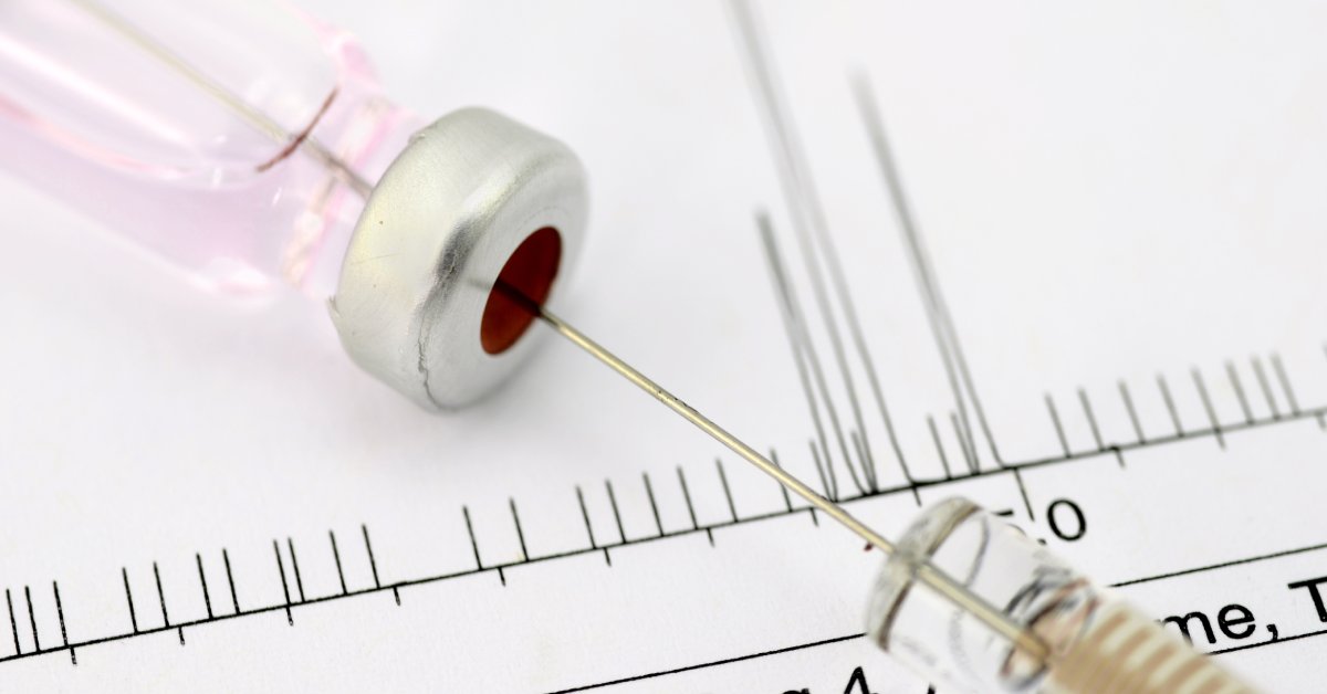 A close-up of the needle of a clear syringe poking a hole in the septa of a lab vial. The vial is on a white surface.