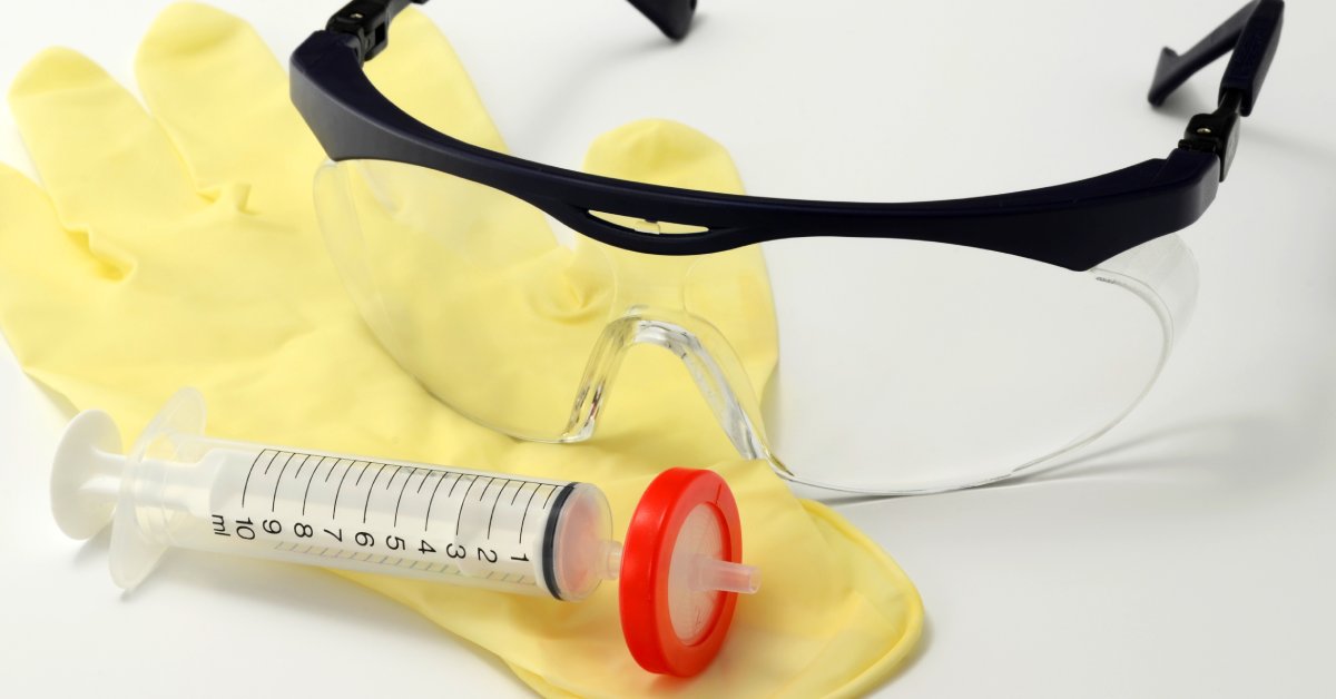 Plastic syringe with a red filter placed on a yellow latex glove next to a pair of safety glasses on top of a white surface.