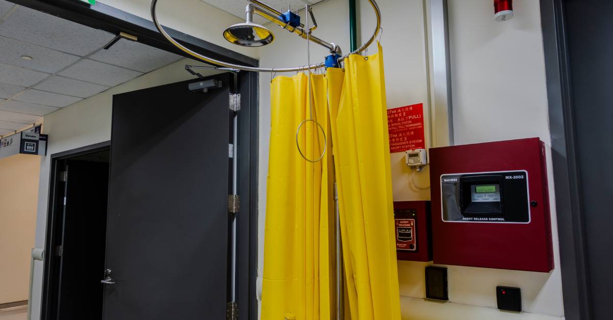 A showerhead surrounded by a yellow curtain next to an exit door and a sink inside a laboratory.