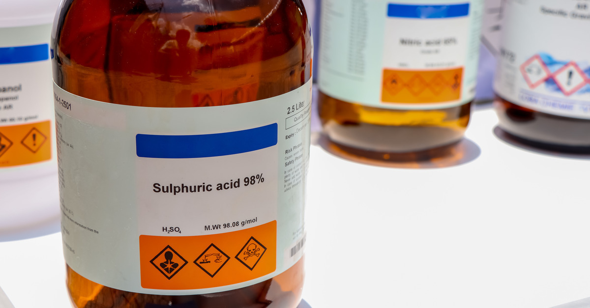 A brown glass bottle with a label displaying the words 'sulphuric acid 98%' placed on a white table with other bottles.