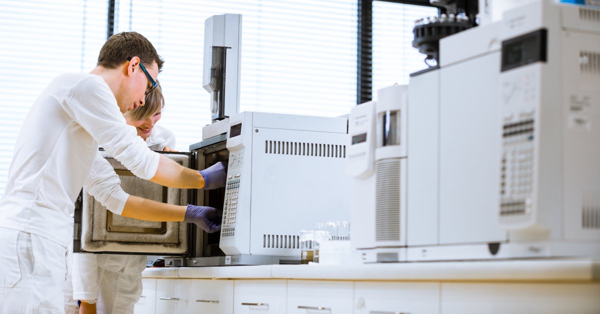 Two scientists wearing lab coats using a large chromatography machine on a counter inside a laboratory.