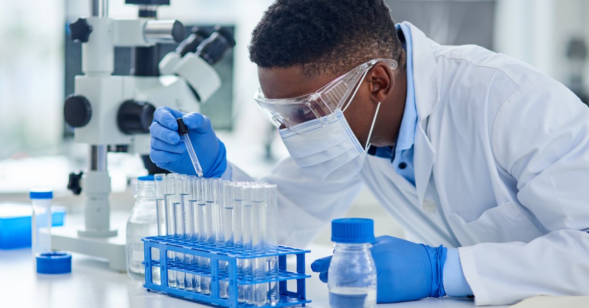 Male scientists wearing goggles, facemask, latex gloves, and white robe while using a dropper on sample tubes.