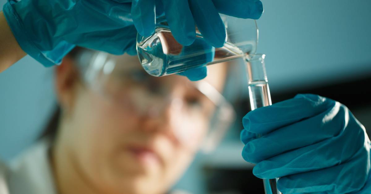 A scientist wearing safety glasses and blue latex gloves, pouring clear water from a beaker into a long glass tube.