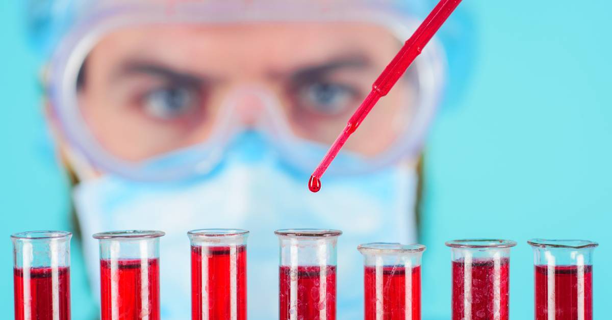 A blurry close-up of a male scientist wearing safety goggles. He's using a pipette full of a red liquid, dripping it into multiple sample tubes.