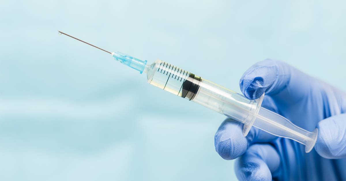 A close-up of a hand wearing a blue latex protective glove, holding a plastic syringe full of a clear liquid.