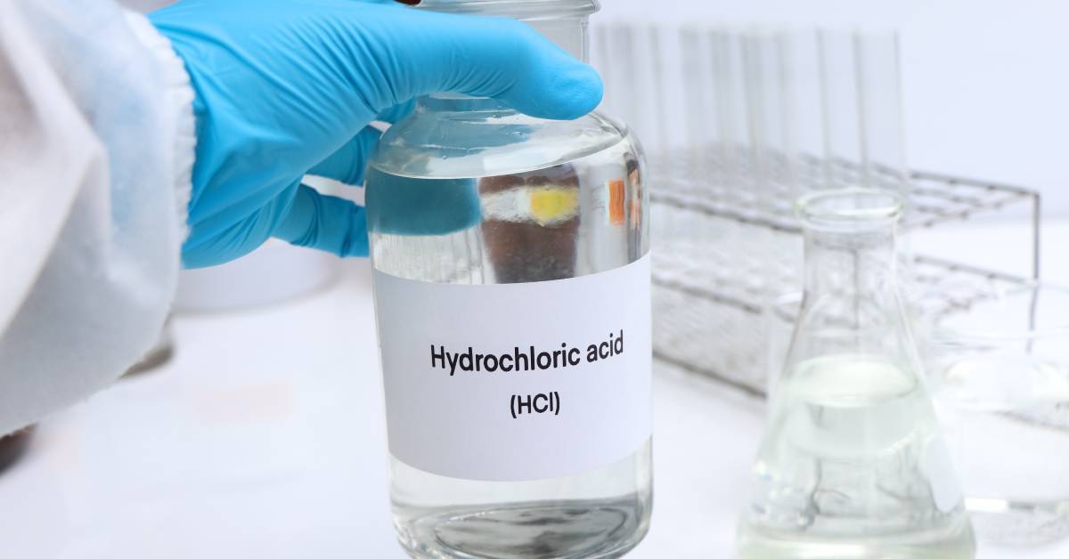 A hand wearing a blue latex glove holds a large glass container full of a clear liquid with a 'HCI' label in a laboratory.