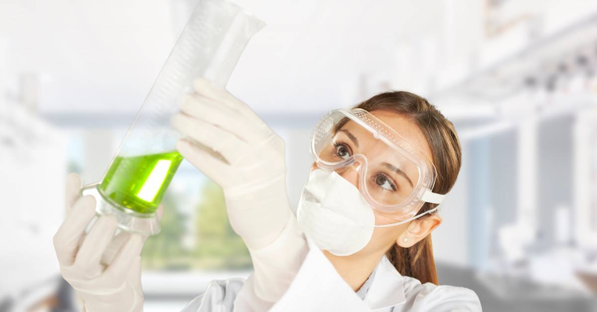 A scientist wearing a white lab coat, latex gloves, safety goggles, and a face mask looks at a container of green liquid.