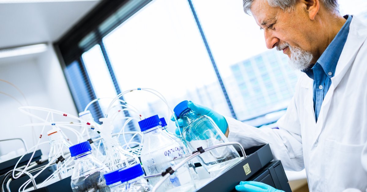 Scientist wearing a face mask, safety goggles, and blue latex gloves holding a vial full of a clear liquid in front of his face.