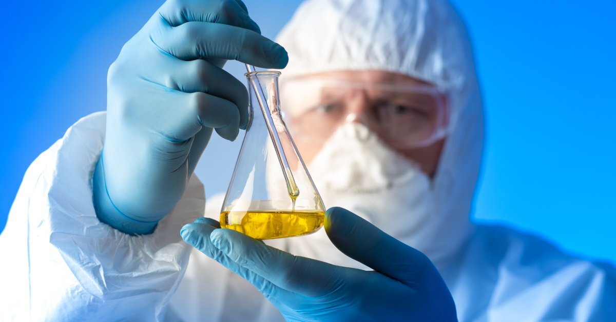 Scientist wearing protective gear holding a large glass container with a viscous yellow liquid and a glass stick.