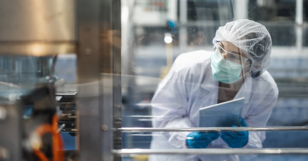 A scientist wearing a mask, safety glasses, a hairnet, and a white robe carrying a tablet while looking at a large lab machine.