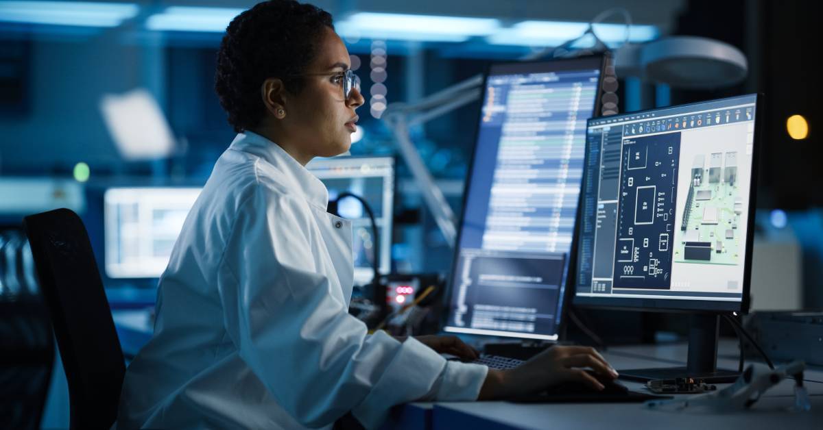 A scientist wearing a white robe and glasses sitting behind a desk looking at two computer screens inside a lab.