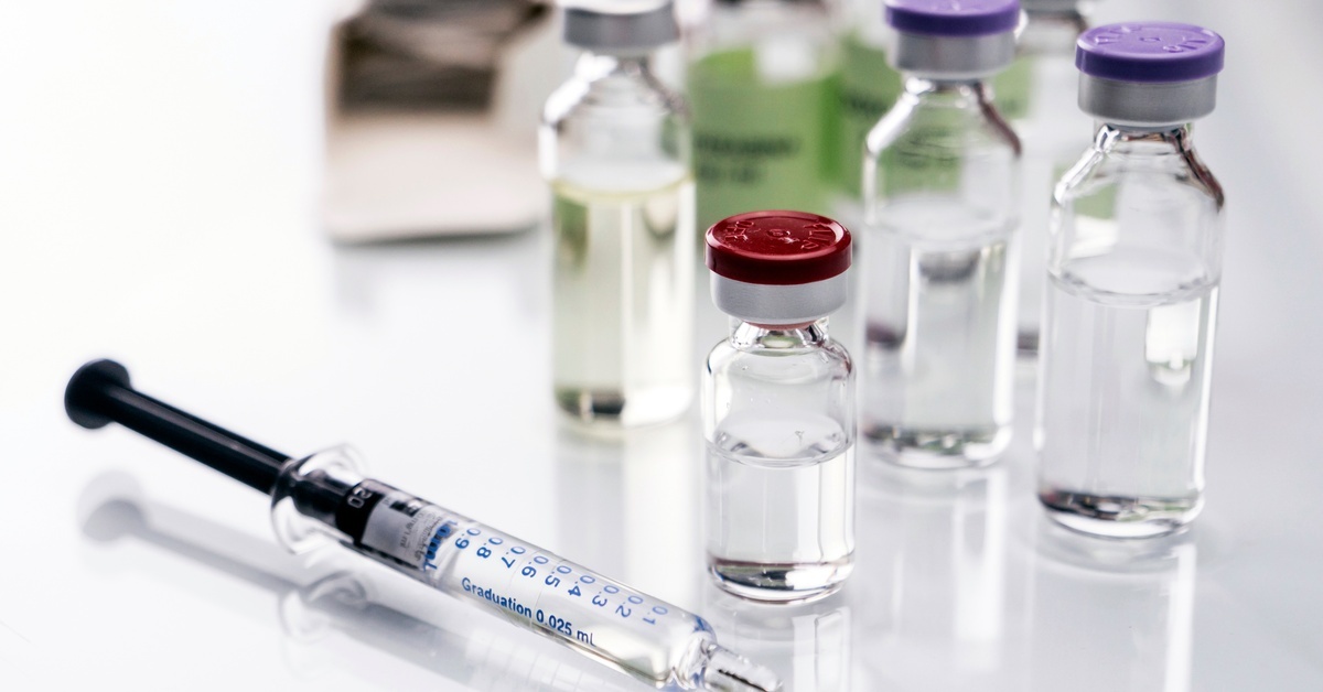 Multiple glass vials of different sizes with caps of various colors are placed on a white surface with a syringe in front.