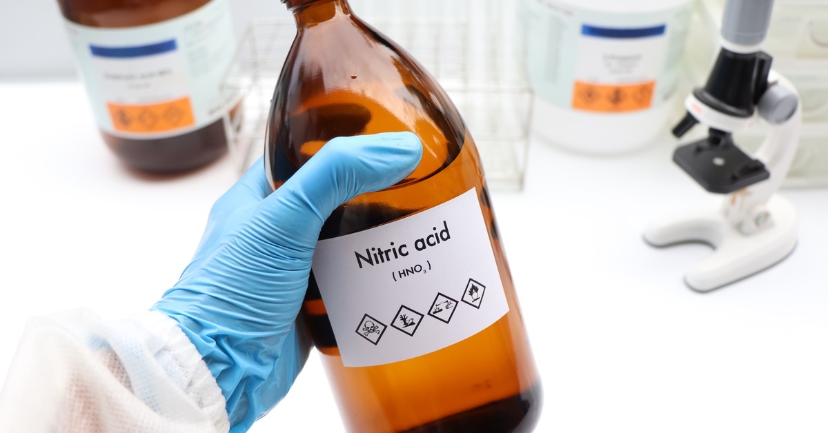 A close-up of a hand wearing a blue latex glove holding an amber glass bottle with a nitric acid label.