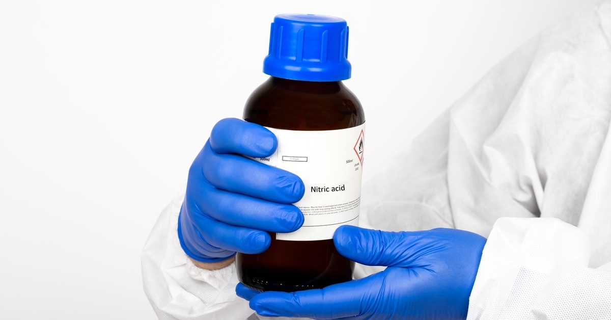 A close-up of a scientist wearing a white lab coat and latex gloves holding a bottle with a nitric acid label.