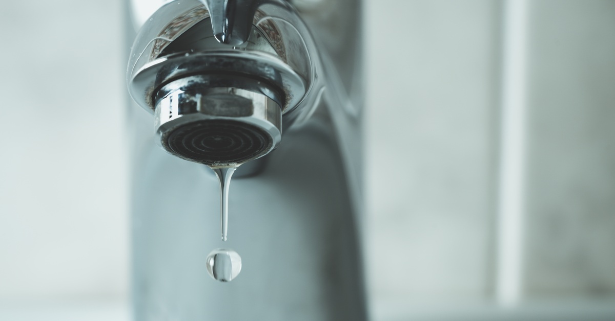A shiny silver water faucet with a droplet hanging from its spout and disconnecting as it falls in a tiled room.